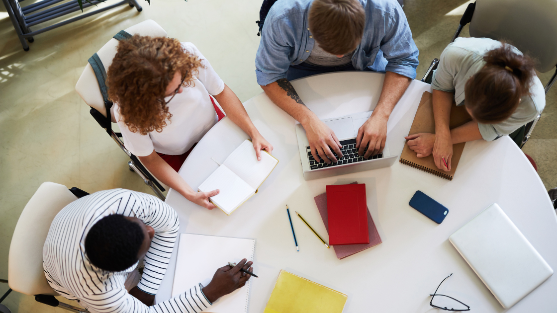 overhead picture of group of four people sitting at a desk with papers and computers in front of them